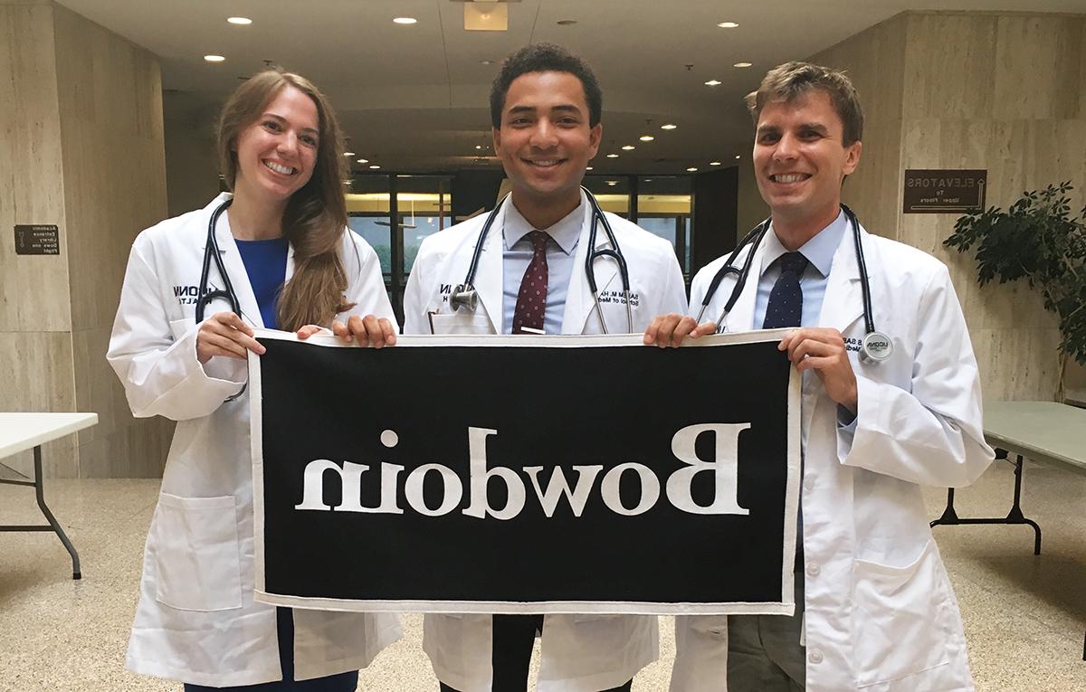 3 students in white medical coats hold up Bowdoin Sign 